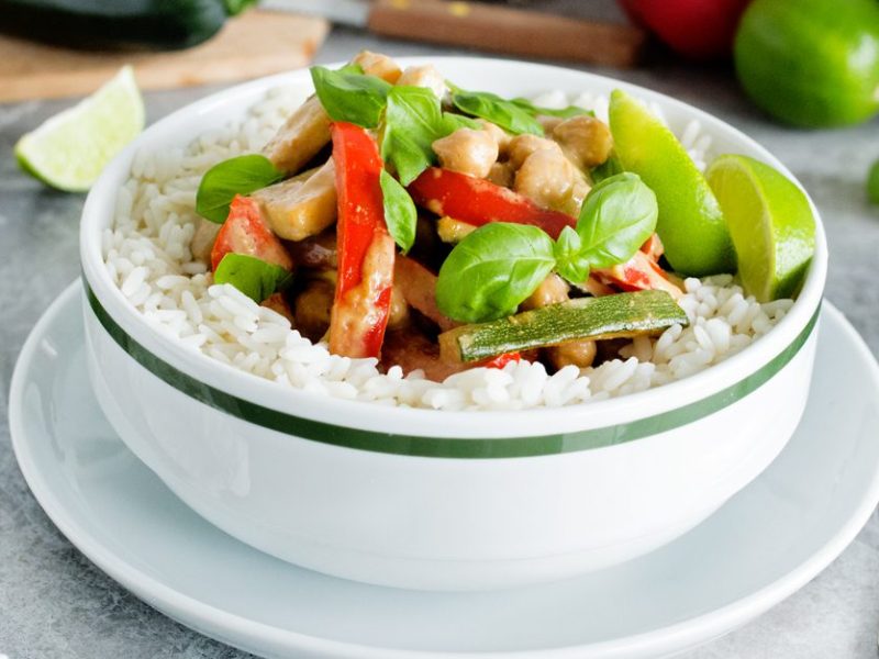 Thai Chickpea-Almond Curry in a white bowl surrounded by fresh limes wedges, basil, and ginger root