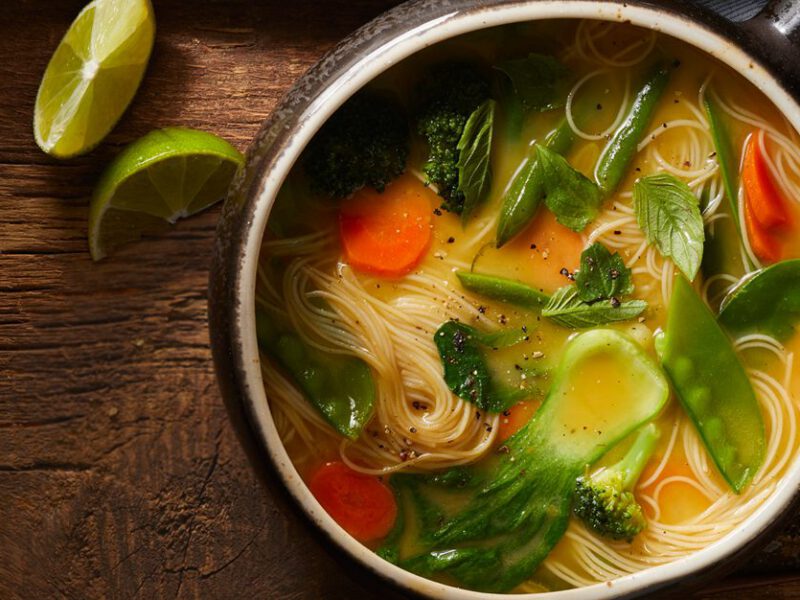 Vegan Thai noodle soup in a rustic pot, with noodles threading through slices of bok choy, carrots, broccoli, and snap peas, garnished with Thai basil, on a wooden table
