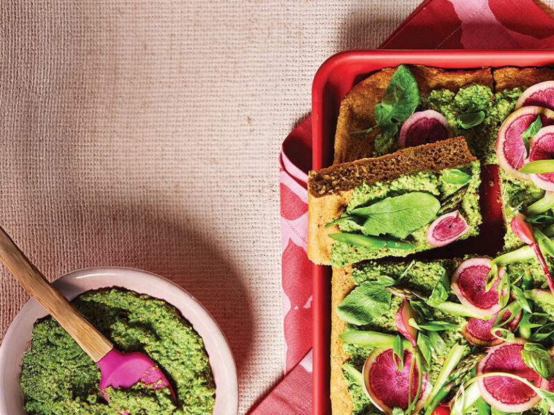 Spring Vegetable Focaccia in a red baking dish, topped witih colorful watermelon radishes and a herbaceous pea spread, next to a small bowl of the green spread