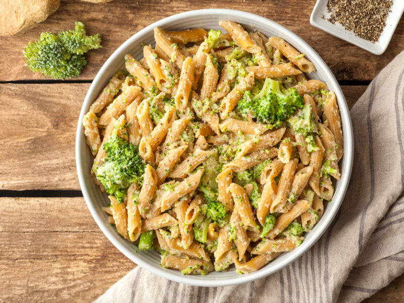Big bowl of vegan celeriac mac and cheese with penne pasta and broccoli florets, on a rustic wooden table with a kitchen towel and seasonings to the side