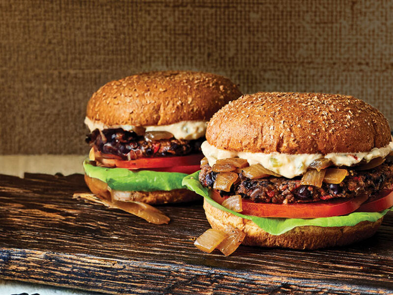 Two Black Bean Smash Burgers with Caramelized Onions on a wooden chopped board with an onion to the left of the board and a small bowl of sauce to the right