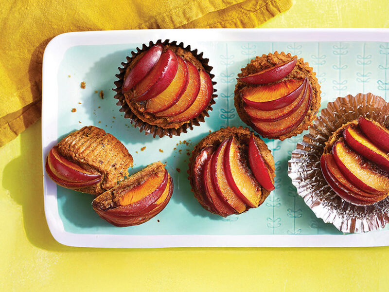 Fresh Plum Muffins in foil muffin papers on a pale blue and white plate with plum slices fanned across the top of each muffin