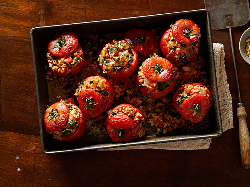 Greek-Style Stuffed Tomatoes on a baking tray with a small bowl of pine nuts to the side