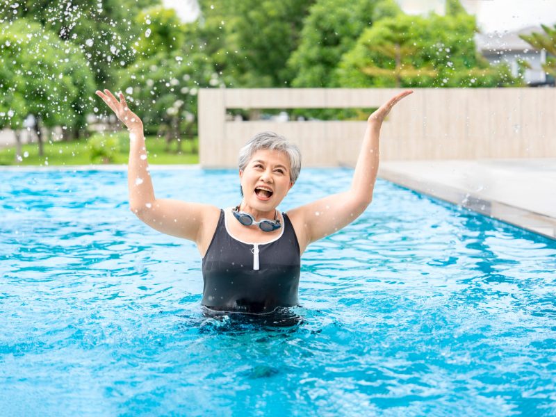A senior woman stands in a pool outside looking joyous with her arms raised upward, wearing an athletic swimsuit and goggles around her neck