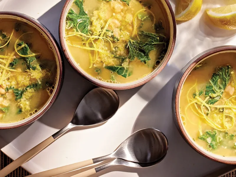Three bowls of Lemon Hummus Soup with Kale on a tray with spoons and lemon wedges on the side