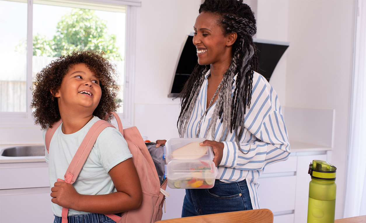 A mother stands behind her daughter in the kitchen ready to put a bag of snacks into her daughters backpack. Both are smiling and happy.